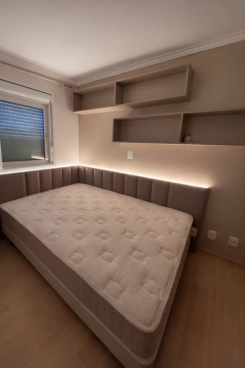 Cama com cabeceira estofada contínua, iluminação indireta e prateleiras em composição na parede