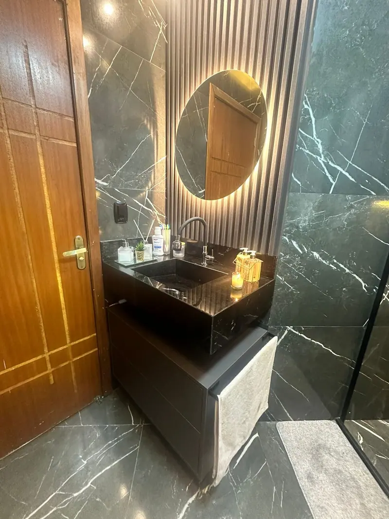 Lavabo com revestimento marmorizado escuro, gabinete preto e espelho redondo iluminado